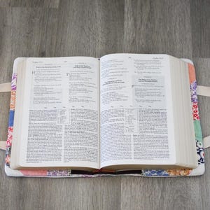 May include: An open book rests in a colorful, patchwork fabric cover with white handles. The cover features a variety of floral and geometric patterns in shades of red, blue, and orange. The book's pages are filled with text.