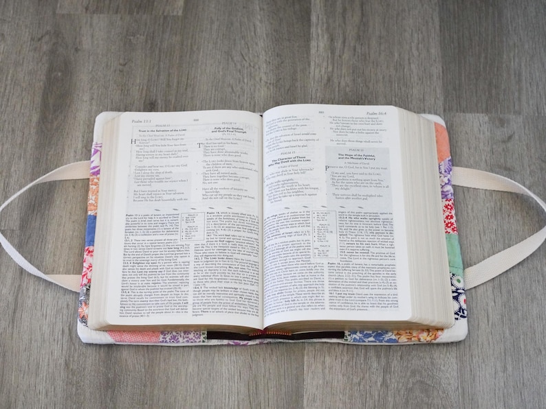 May include: An open book rests in a colorful, patchwork fabric cover with white handles. The cover features a variety of floral and geometric patterns in shades of red, blue, and orange. The book's pages are filled with text.
