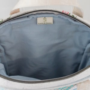 May include: Open view of a floral-patterned handbag with a light blue interior. The bag has a zipper closure and a cream-colored strap. The bag's exterior features a white background with pink and green floral designs. The label inside the bag reads "PLAINS & PRAIRIE".