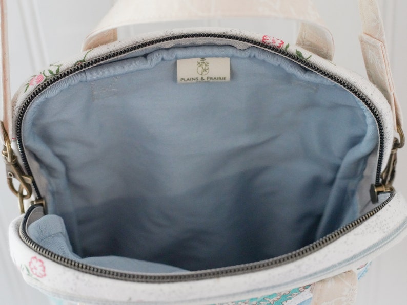 May include: Open view of a floral-patterned handbag with a light blue interior. The bag has a zipper closure and a cream-colored strap. The bag's exterior features a white background with pink and green floral designs. The label inside the bag reads "PLAINS & PRAIRIE".