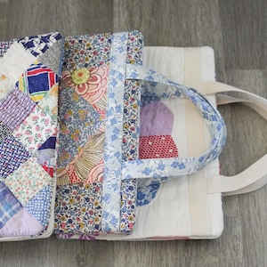 May include: A patchwork tote bag with a variety of colorful fabric patterns. The bag features a handle made of blue floral fabric and a white canvas strap. The bag is open, revealing the interior with more patchwork and a white lining.