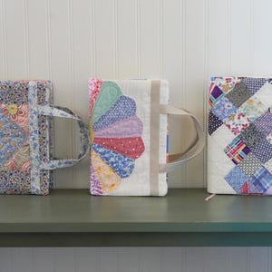 May include: Three quilted book covers with handles. The covers feature floral and patchwork designs in various colors, including blue, pink, and purple. They are displayed on a green surface against a white wall.