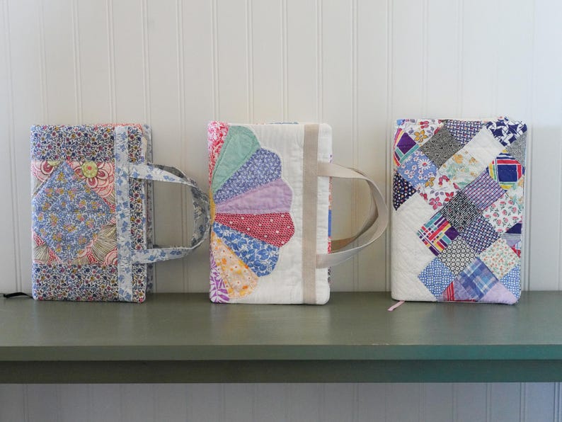 May include: Three quilted book covers with handles. The covers feature floral and patchwork designs in various colors, including blue, pink, and purple. They are displayed on a green surface against a white wall.