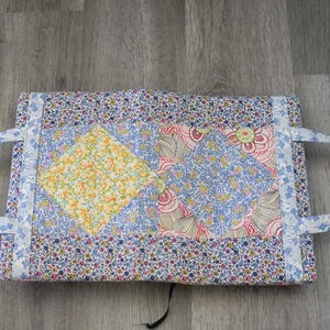 May include: A handmade fabric tote bag with floral patterns in blue, pink, and yellow. The bag features a patchwork design with a yellow diamond shape in the center. It has two handles and a button closure. The bag is approximately 12 inches wide and 9 inches tall.