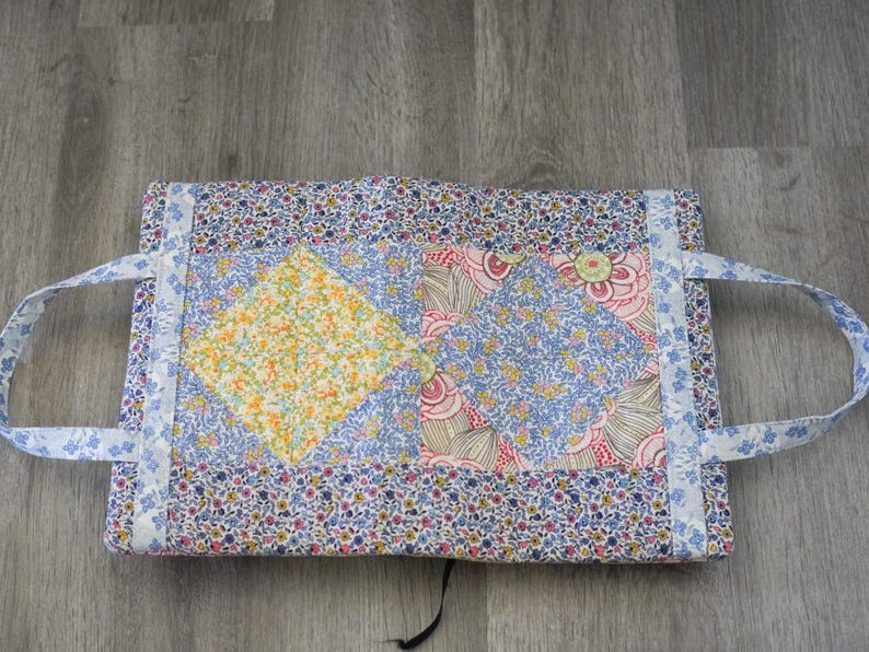 May include: A handmade fabric tote bag with floral patterns in blue, pink, and yellow. The bag features a patchwork design with a yellow diamond shape in the center. It has two handles and a button closure. The bag is approximately 12 inches wide and 9 inches tall.