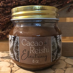 May include: A glass jar with a gold lid filled with a brown substance. The label reads "Cacao + Reishi Electuary" and "8 oz". The jar is set on a wooden surface, with dried plants in the background.