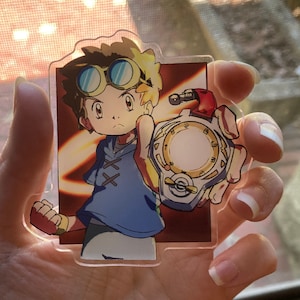 Peut inclure: Un porte-clés en acrylique transparent représentant un personnage de dessin animé aux cheveux blonds hérissés portant des lunettes de protection et une chemise bleue. Le personnage tient un appareil doré et argenté avec un cadran circulaire.