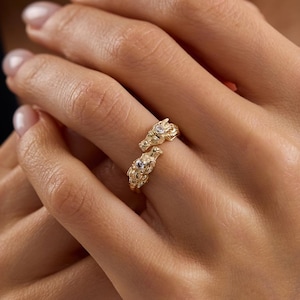 May include: A gold-colored ring with a unique design, featuring two ornate, textured sections that appear to wrap around the finger. Small, clear gemstones are set into the ring, adding sparkle. The ring is displayed on a person's hand.