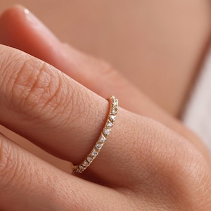May include: A delicate gold ring with a continuous row of small, square-cut gemstones. The ring is worn on a finger, showcasing its elegant design and sparkle. The ring's band is thin, emphasizing the gemstones' brilliance.