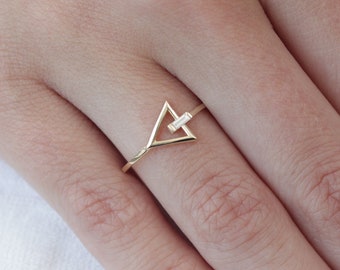 Op de afbeelding: Een gouden driehoekring met een kleine diamant in het midden. De ring zit op een vinger met lichtroze nagellak.