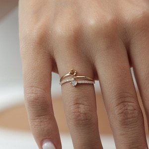 Peut inclure: Une bague en or avec une petite pierre transparente et une bande délicate. La bague est portée à l'annulaire d'une main avec de longues ongles blancs.