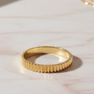 May include: A gold ring featuring a vertical ribbed pattern. The ring sits on a marble surface, with a gold necklace and a dried leaf in the background. The ring is a simple band with a textured surface.