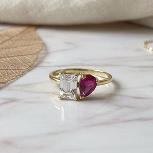 May include: A gold ring featuring a rectangular, clear gemstone and a pear-shaped, red gemstone. The ring is set against a marble surface with a gold chain and a leaf in the background. The ring is a piece of jewelry.