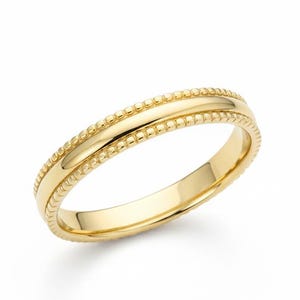 May include: A gold ring with a beaded edge design. The ring has a smooth, polished center band and a delicate, textured border. The ring is set against a white background, highlighting its bright, shiny appearance. The ring is a classic piece of jewelry.