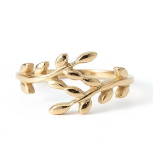 May include: A gold-colored ring featuring a delicate leaf design. The ring's band is thin, and the leaves wrap around the finger, creating an elegant and natural look. The ring is set against a white background.