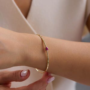 May include: A gold-colored bangle bracelet with a small, oval-shaped, red gemstone. The bracelet has a delicate, textured design and is worn on a wrist. The jewelry is a simple, elegant accessory.