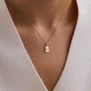 May include: A gold-colored necklace with a rectangular pendant. The pendant has a sunburst design around a central letter. The necklace is worn against a white top.