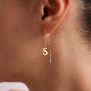 May include: A gold-coloured earring featuring the letter "S". The earring is a delicate, drop-style design, with the letter suspended from a thin chain. The earring is shown in wear, with the ear and part of the neck visible.