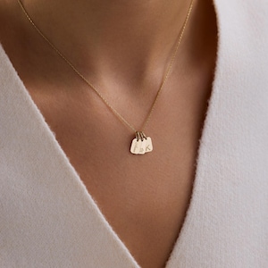 May include: A gold-colored necklace with three small, rectangular pendants. Each pendant has a rounded top and is engraved with a single initial or a short name. The necklace is worn against a white garment.