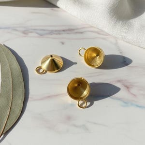 Könnte beinhalten: Drei kleine, goldfarbene Metalltrichter mit einem Ring an der Seite. Ein Trichter ist umgedreht und zeigt das spitz zulaufende Ende. Die anderen beiden Trichter stehen aufrecht, mit einem abgerundeten Boden und einer kleinen Öffnung oben. Die Trichter liegen auf einer Marmoroberfläche.