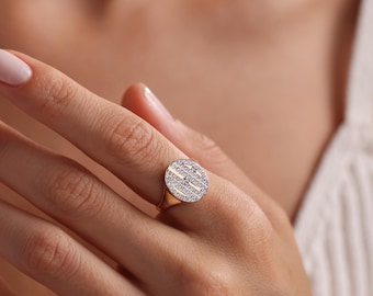 May include: A gold ring with a round face, set with small, sparkling stones in a geometric pattern. The ring is on a finger, showcasing the design and the warm tones of the metal and gems. The ring is a piece of jewelry.