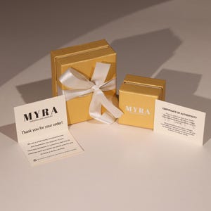 May include: A gold-colored gift box with a white ribbon bow, a smaller box, and two cards. The cards read "MYRA Personalized Jewelry" and "Thank you for your order!" and "Certificate of Authenticity".