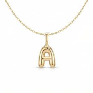 以下が含まれることがあります： 金色のペンダントネックレスで、文字「A」が特徴です。 ペンダントは丸みを帯びたバブルのようなデザインで、繊細なチェーンから吊り下げられています。 ネックレスは白い背景に表示されています。