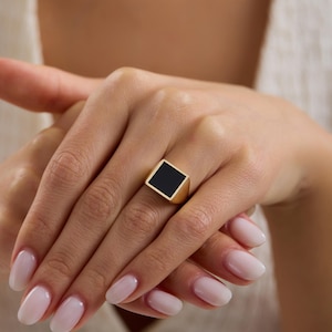 May include: A gold signet ring featuring a flat, square-shaped black inlay centered on the band. The ring has a polished metallic finish and a geometric design, with the square face positioned prominently on the finger for a clean, bold look.