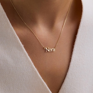 May include: A gold necklace with the name "Nora" as a pendant. The necklace is suspended from a fine chain. The jewellery is a simple, elegant piece. The chain is visible against the person's skin.