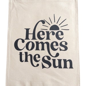 以下が含まれることがあります： ベージュのキャンバス製の壁掛けタペストリーで、黒い文字で「Here Comes the Sun」と書いてあり、スタイリッシュな黒い太陽のグラフィックが描かれています。