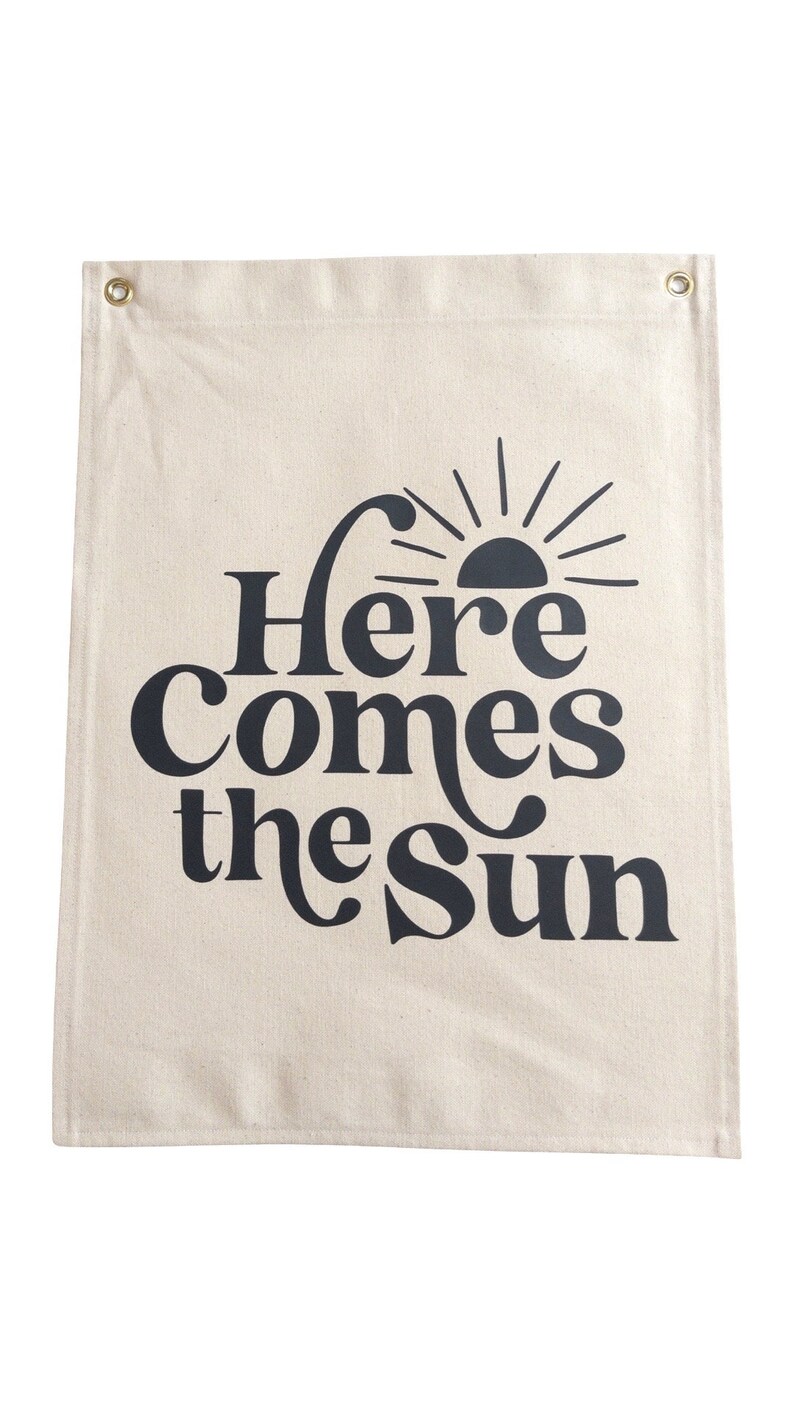 以下が含まれることがあります： ベージュのキャンバス製の壁掛けタペストリーで、黒い文字で「Here Comes the Sun」と書いてあり、スタイリッシュな黒い太陽のグラフィックが描かれています。