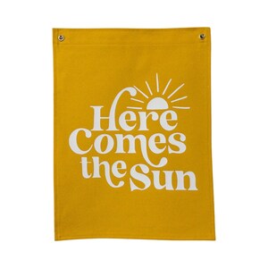以下が含まれることがあります： 「Here Comes the Sun」という白い文字とスタイリッシュな太陽のグラフィックが描かれた黄色のキャンバスバナーです。