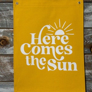 以下が含まれることがあります： 黄色いキャンバスの横断幕に、白い文字で「Here Comes the Sun」と書いてあり、スタイリッシュな太陽のグラフィックが描かれていて、光線が放射されています。