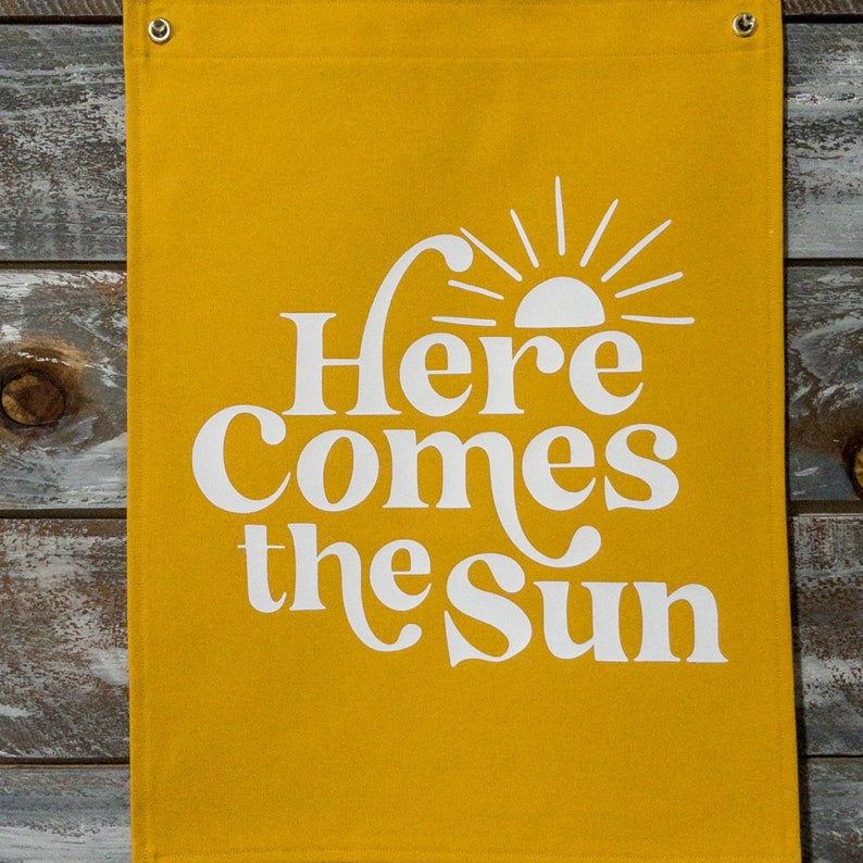 以下が含まれることがあります： 黄色いキャンバスの横断幕に、白い文字で「Here Comes the Sun」と書いてあり、スタイリッシュな太陽のグラフィックが描かれていて、光線が放射されています。