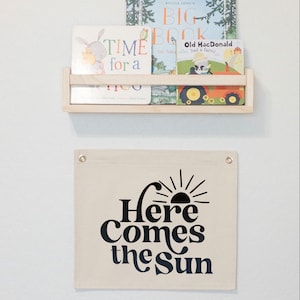 以下が含まれることがあります： ベージュのキャンバスバナーに、黒い文字で「Here Comes the Sun」と書いてあり、その上にスタイリッシュな太陽のグラフィックがあります。
