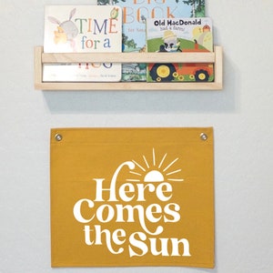以下が含まれることがあります： 白い文字で「Here Comes the Sun」と書かれた黄色のキャンバスバナー。スタイリッシュな太陽と光線が描かれています。バナーの上部には銀色のグロメットが2つ付いています。