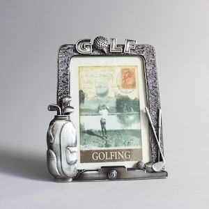 Könnte beinhalten: Silberfarbener Bilderrahmen mit Golf-Motiv. Der Rahmen zeigt eine Golftasche, Golfschläger und das Wort "GOLF" mit einem Golfball. Der Rahmen enthält eine Vintage-Postkarte mit einem Golfer auf einem Golfplatz.