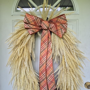 May include: A decorative wreath featuring a circular base of brown twigs adorned with long, pale yellow, feathery foliage. A large, multi-looped bow made of orange, brown, and cream plaid fabric is centered at the top, with two long tails hanging down.