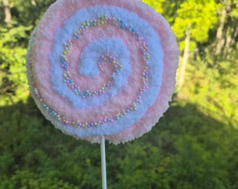 May include: A large, decorative lollipop with a pink, white, and blue swirl design. The lollipop is adorned with small, colorful sprinkles and is held by a white stick. The background is a blurred view of green foliage.