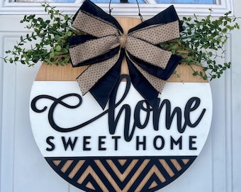 May include: Round wooden door sign with the words "Home Sweet Home" in black script. The sign has a white and wood grain background, a black geometric design at the bottom, and a large bow with black and tan polka dot ribbon.