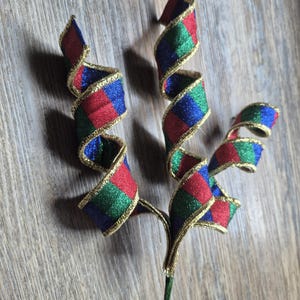May include: A decorative floral pick featuring three spiraled ribbons with a checkered pattern in red, green, and blue. Each ribbon is edged with metallic gold trim, and the stems are joined together at the base into a single green wire.