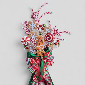 May include: A festive Christmas decoration featuring gingerbread men, candy canes, and snowflake ornaments. The arrangement includes a large red and green bow, creating a cheerful holiday display. The color palette includes red, green, white, and brown.