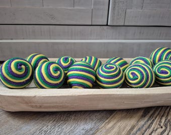 May include: A collection of decorative balls in a wooden tray. The balls are adorned with a spiral design in yellow, green, and purple. The tray sits on a wooden surface, with a neutral-toned background.