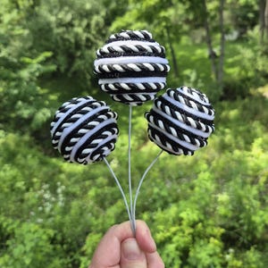 May include: Three decorative spheres with a black and white rope design, held by a person. The spheres are on thin, gray stems, with a backdrop of green foliage.