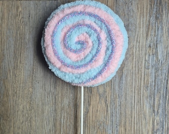 May include: A whimsical, oversized lollipop with a spiral design. The candy features alternating bands of pastel pink and blue, with a shimmering glitter spiral. The lollipop is mounted on a white stick, creating a playful, decorative item.