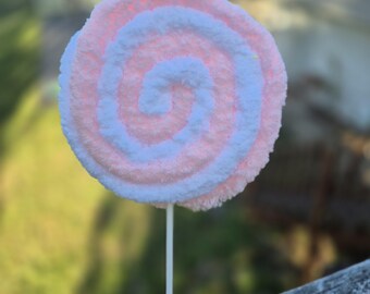 May include: A large, whimsical lollipop with a spiral design. The candy features alternating bands of soft pink and white, creating a fluffy, textured appearance. It is mounted on a white stick, suggesting a playful, decorative item.