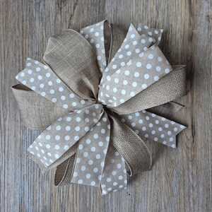 May include: A decorative bow made of tan burlap and a ribbon with white polka dots. The bow is tied in a full, multi-loop style, with several layers of ribbon creating a textured appearance. The bow is set against a wooden background.