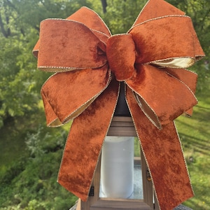 May include: A large, decorative bow made of burnt orange velvet ribbon with gold metallic edges. The bow is tied in a multi-loop style with two long, hanging tails and is positioned atop a wooden lantern containing a white pillar candle.