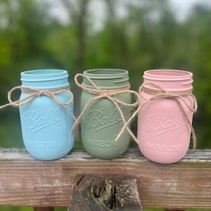 May include: Three mason jars painted in light blue, green, and pink with a brown twine bow around the top of each jar. The jars are labeled "Ball Mason" on the front.
