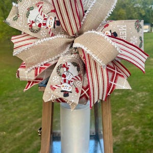 May include: A wooden lantern with a white candle inside. The lantern is decorated with a large burlap and red and white striped bow with snowman and Christmas themed fabric accents. The bow has the words "Merry Christmas" printed on it.